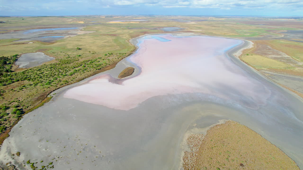 Aerial cinematic view over a salt lake with soft pastel pink colours visible across the surface. Smooth and cracked textures form abstract patterns, creating a calm and visually striking landscape