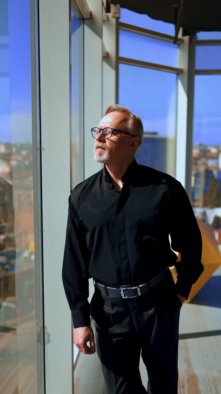 Adult man stands leaning to the window looking outside on sunny day. Businessman wearing black shirt and trousers puts off glasses. Vertical video.