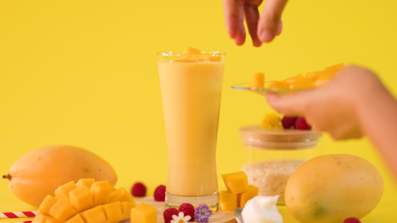Human hand adds fresh mango pieces onto tall tropical smoothie against bright yellow background