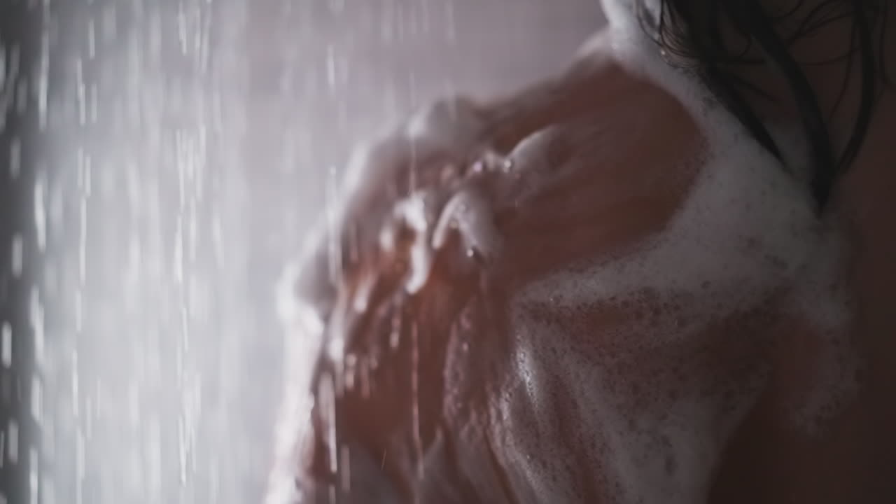 el agua lava la espuma del hombro de la mujer en el baño de cerca. la dama toma una ducha caliente y limpia el cuerpo con espuma de jabón en el baño. relajación en el spa