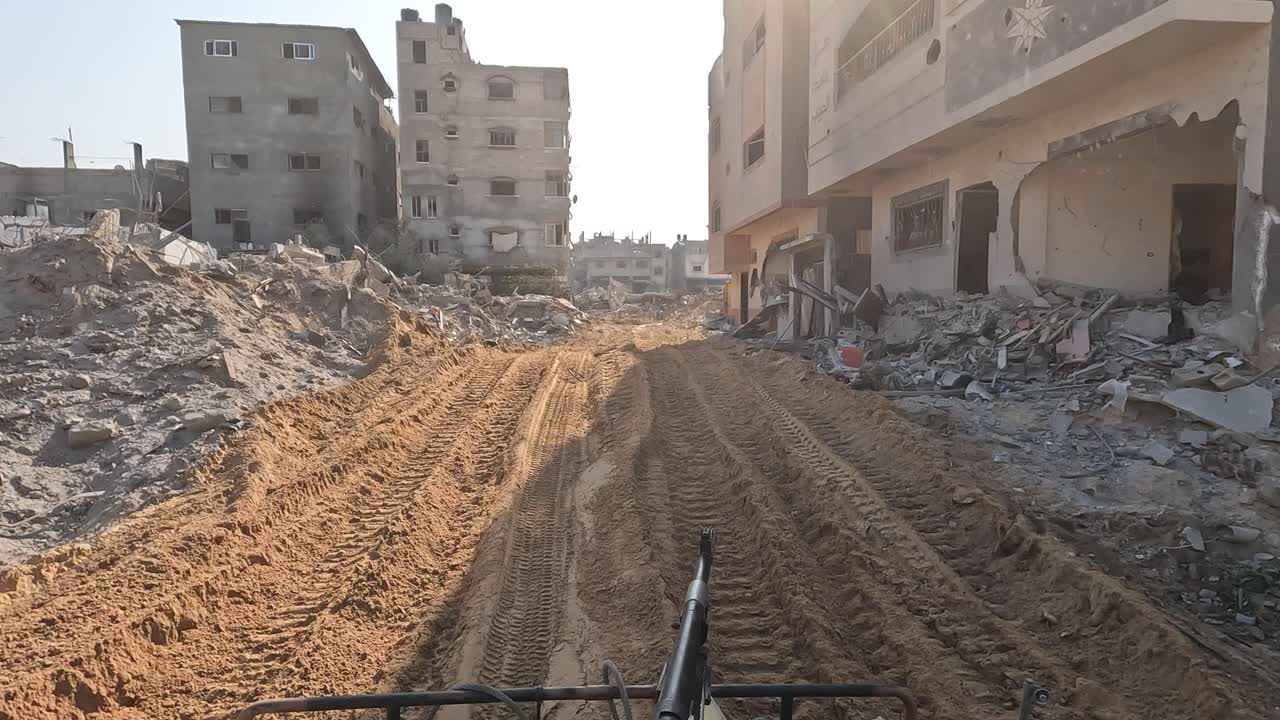Striscia di Gaza — POV da veicolo militare che percorre una strada sterrata fangosa, fiancheggiata da edifici pesantemente danneggiati e detriti in un'area urbana devastata.