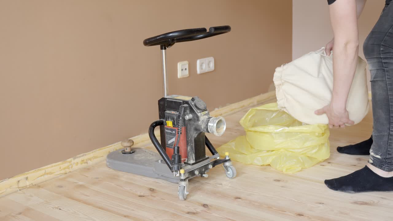 Person sanding wooden floor edges, using a machine with a cloth bag to collect dust