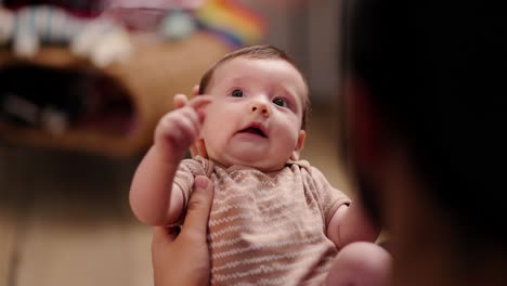 Top-view-of-a-little-baby-girl-in-light-brown-with-her-young-brunette-father-in-a-modern-apartment.-Over-the-shoulder-of-a-happy-young-brunette-man-holding-a-little-baby-girl-in-his-arms