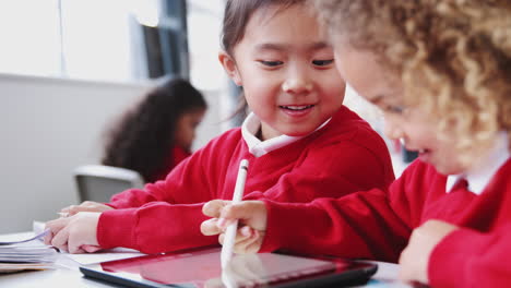 two infant school girls drawing with tablet computer and stylus at a desk in class, close up