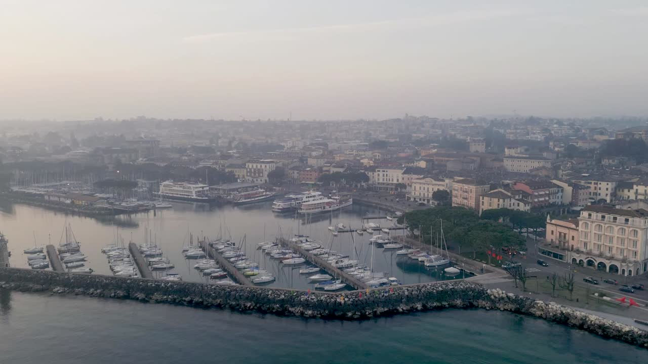 Hyperlapse time lapse drone flight lake garda near town desenzano del garda in the morning, fog hangs over lake and harbor with boats, road traffic rushes by.