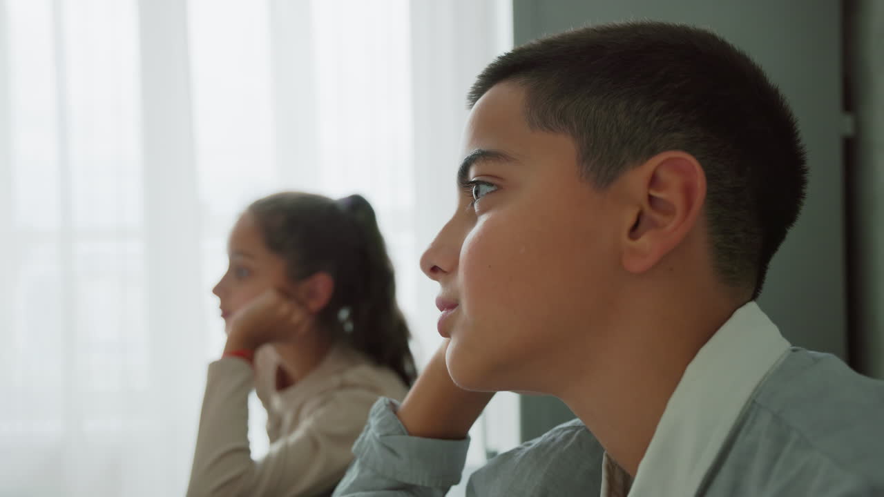 boy and girl in school uniforms sitting at table quietly in morning light near window with pensive expressions waiting indoors during calm home routine moment surrounded by soft neutral tones