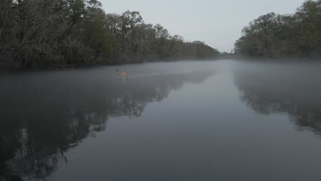 Hombre-Barbudo-Con-Un-Gato-Amarillo-Remando-En-Un-Kayak-Desde-Una-Espesa-Niebla-Que-Cubre-El-Río