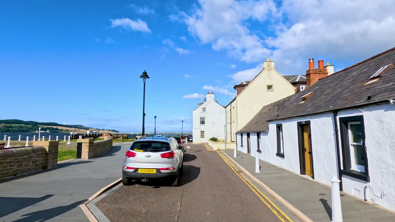 Car drives along sunlit, narrow street with historic cottages toward waterfront in coastal Scotland