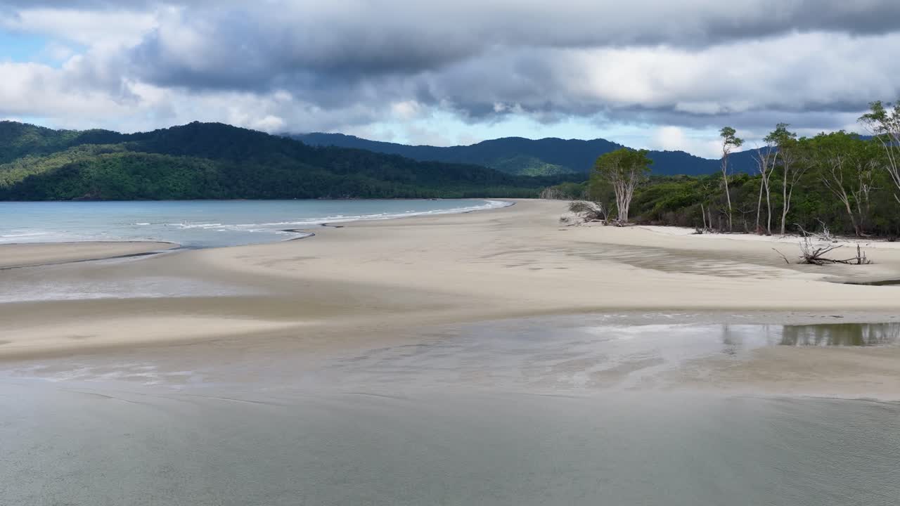 Aerial footage glides over a deserted sandy beach bordered by lush rainforest and calm water, under dramatic cloudy skies with soft natural lighting