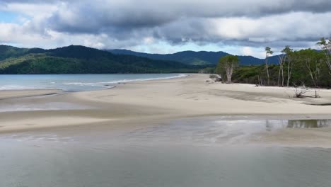 aerial footage glides over a deserted sandy beach bordered by lush rainforest and calm water, under dramatic cloudy skies with soft natural lighting