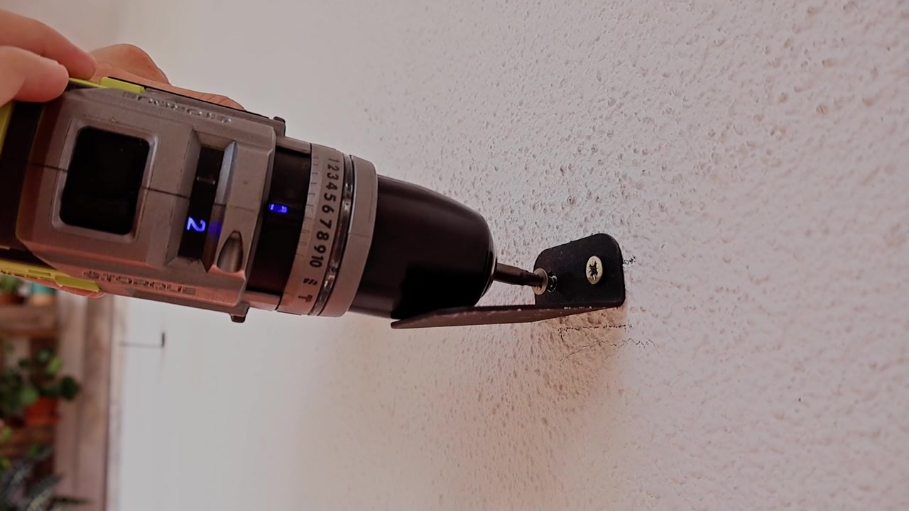Hand drills into wall with metal bracket. Drill bit spins into textured surface firmly. Wall mount holds steady after securing. DIY scene shows precise, confident work. Use for home improvement