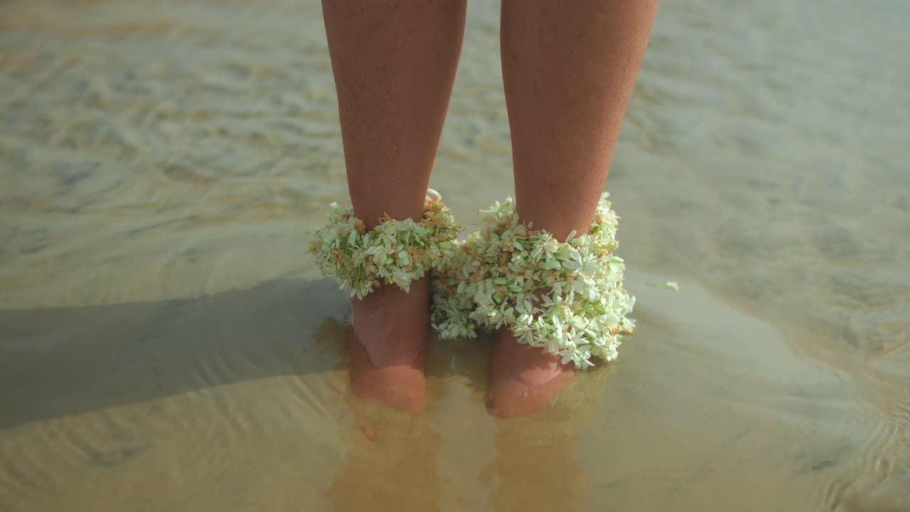 Reverse tracking shot showing a woman’s feet adorned with natural flower anklets, standing in shallow sandy and muddy water on a sunny day