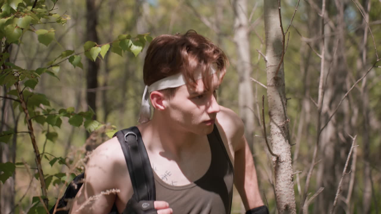 young man navigating dense forest path dodging branches and foliage while walking boldly through undergrowth wearing headband tank top gloves and backpack sunlight filtering through trees