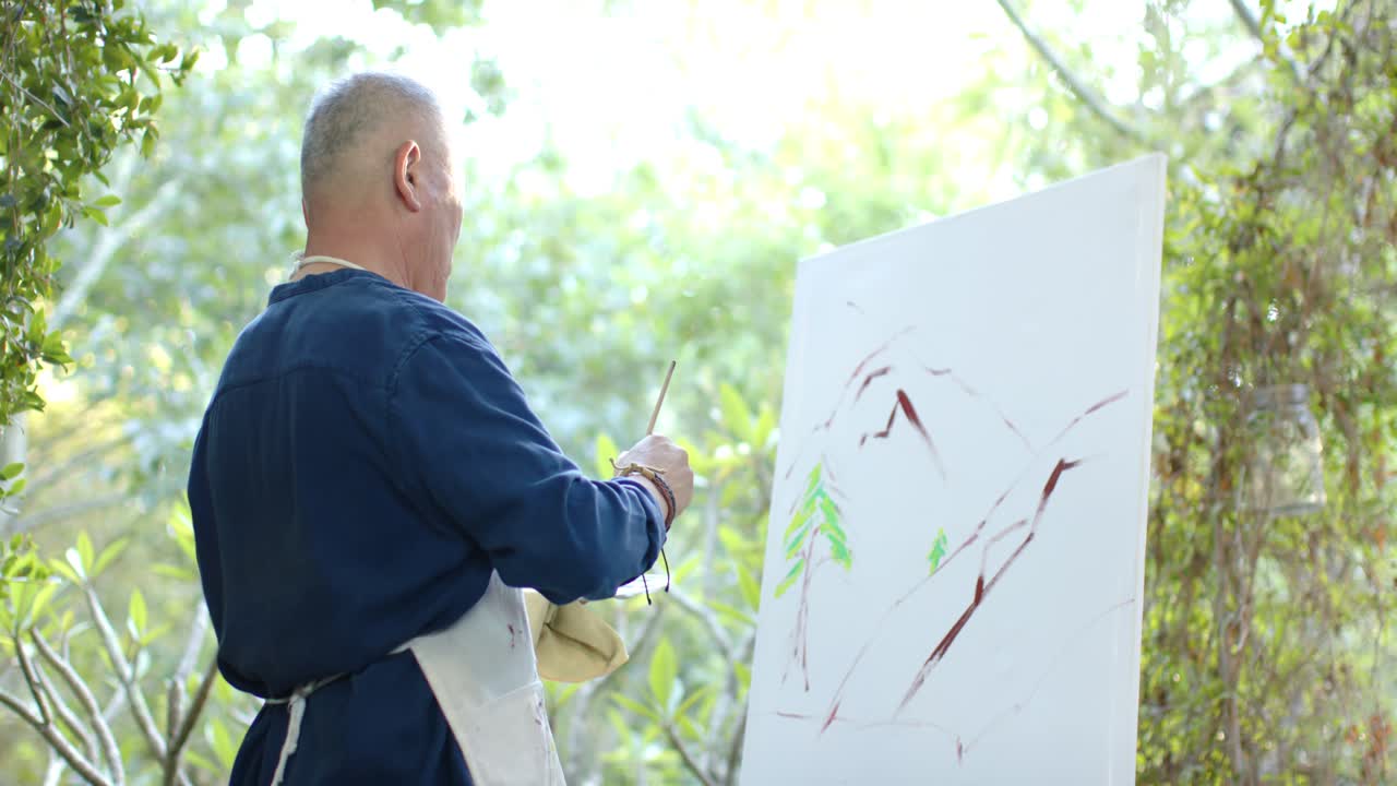 Pittore anziano asiatico con un grembiule che studia la tela sul cavalletto, dipingendo linee marroni per rifinire i dettagli.