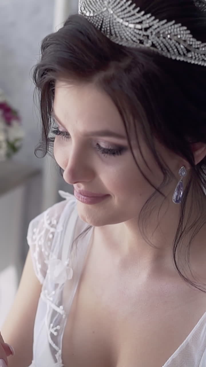 young pretty bride in white lacy dress with wonderful tiara on dark hairstyle smiles against flowers in room slow motion