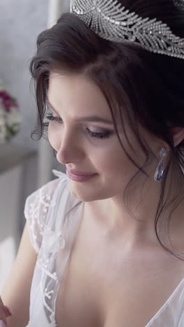 young pretty bride in white lacy dress with wonderful tiara on dark hairstyle smiles against flowers in room slow motion