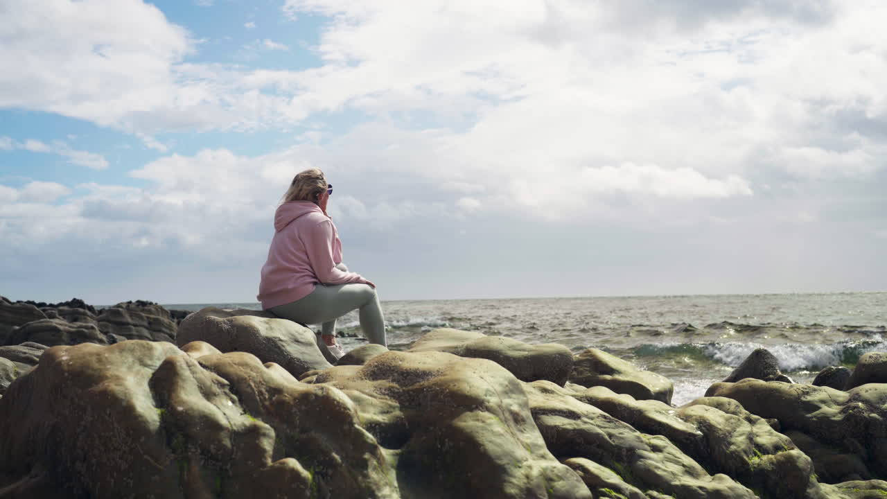 Handheld shot of blond caucasian woman sitting on rocks in front of the sea in Scotland, United Kingdom