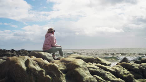 handheld shot of blond caucasian woman sitting on rocks in front of the sea in scotland, united kingdom