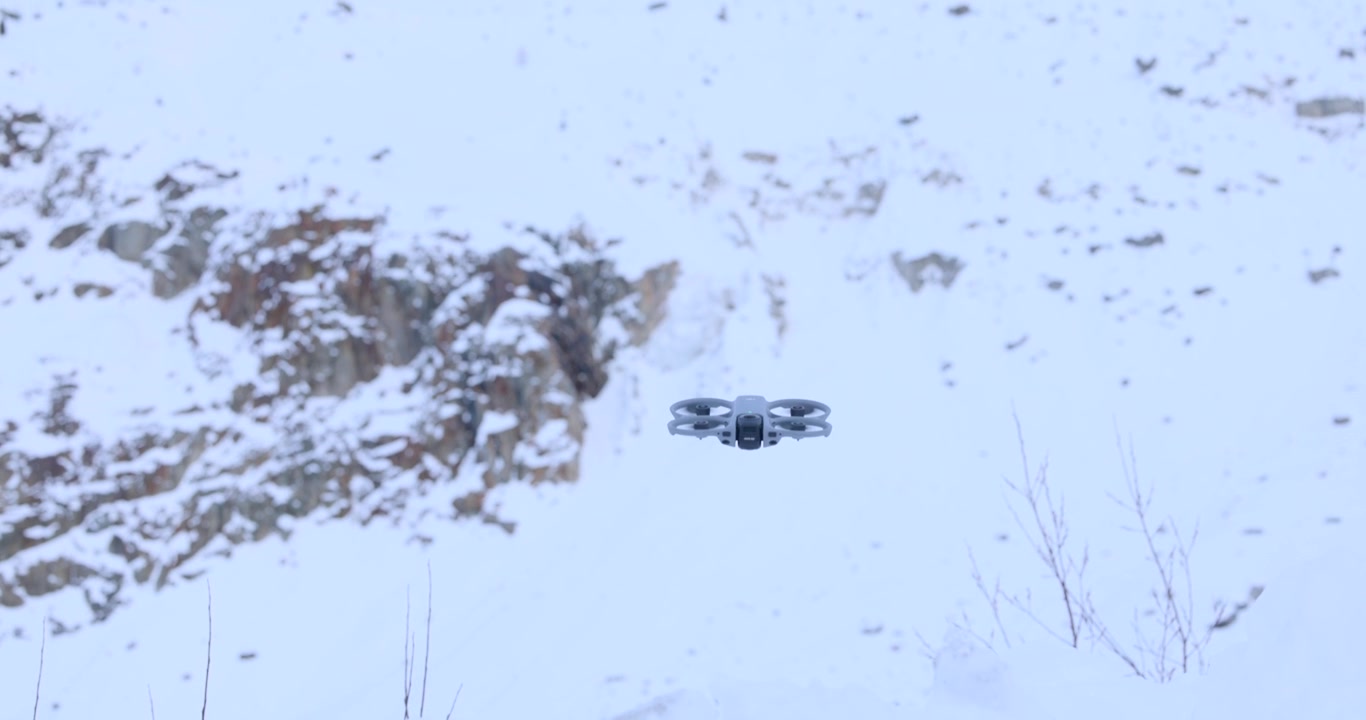 Close up of a compact FPV drone hovering in mid-air against a backdrop of snow covered mountains in winter