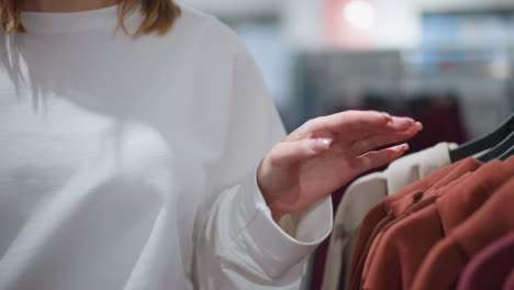 close-up of woman browsing clothing rack, gently touching and observing beige and brown garments, focus on hand interacting with fabric