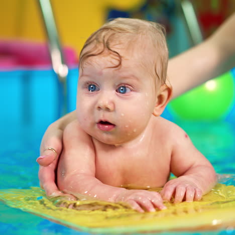 beautiful blue-eyed caucasian baby lies on the board in the swimming pool. woman is moving the kid by the pool.
