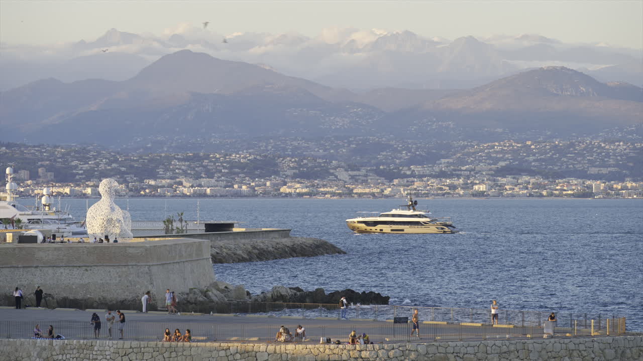 Antibes, France - June 8, 2025: People walking on the coast of the city with The Nomad by Jaume Plensa sculpture on the background