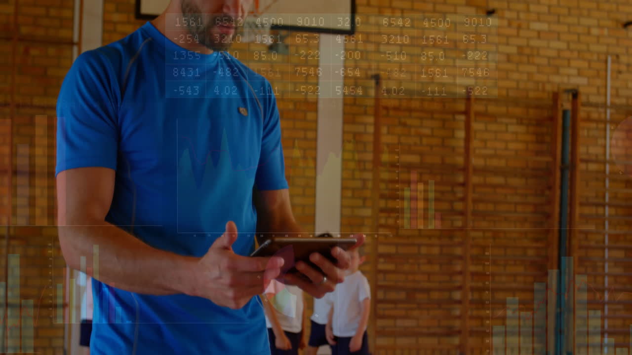 Mid adult coach holding tablet standing in school gym using data tech with digital graphs floating