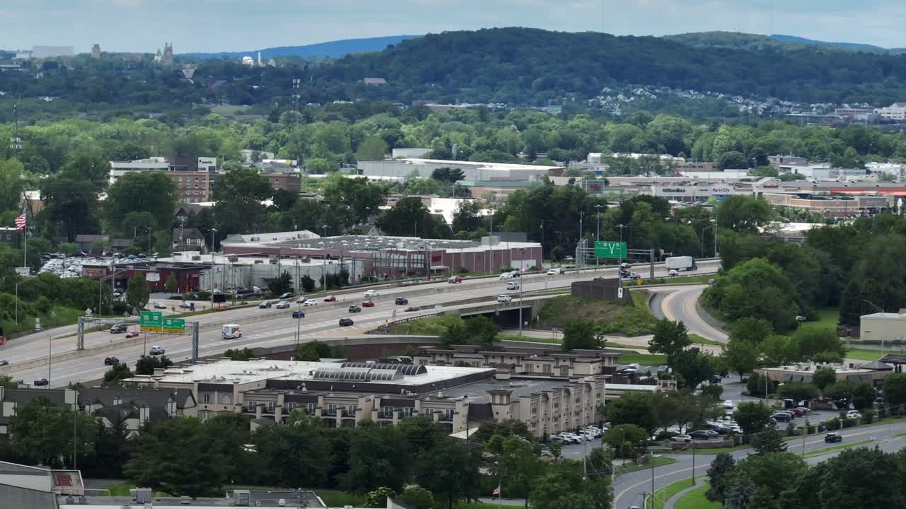 American highway traffic in suburb of town. Aerial wide shot. Green trees in suburb neighborhood and warehouses of company. Green mountain trees in distance.