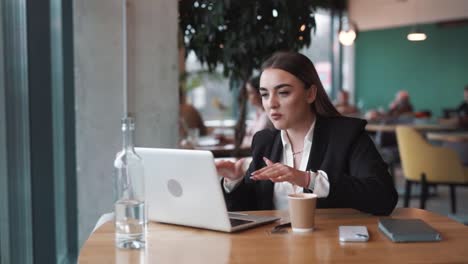 Una-Mujer-Seria-Sentada-En-Un-Elegante-Café-Vestida-De-Negocios-Con-Una-Computadora-Portátil,-Hablando-En-Una-Videollamada,-Discutiendo-Furiosamente-Y-Haciendo-Gestos-Con-Las-Manos