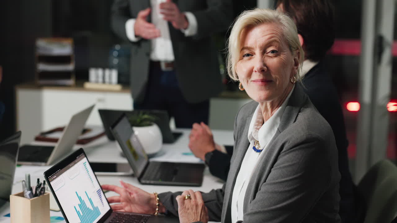 Donna d'affari matura che lavora al laptop e sorride durante una riunione d'ufficio