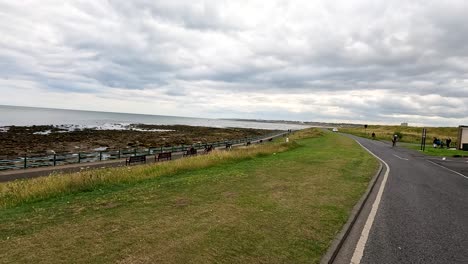 several cyclists travel along a curving coastal road beside a rocky shoreline under overcast skies, with steady camera movement and natural daylight