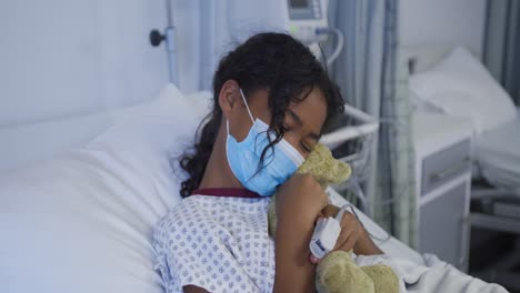 feliz chica de raza mixta acostada en una cama de hospital con una máscara de cara abrazando un oso de peluche