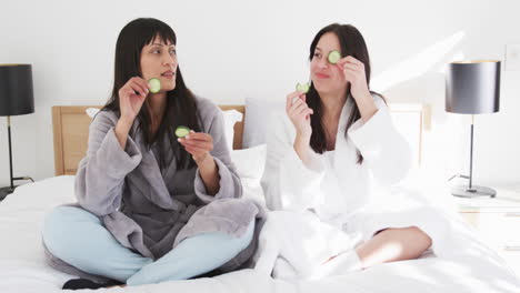 happy biracial mother and adult daughter in robes putting cucumber slices on eyes, slow motion