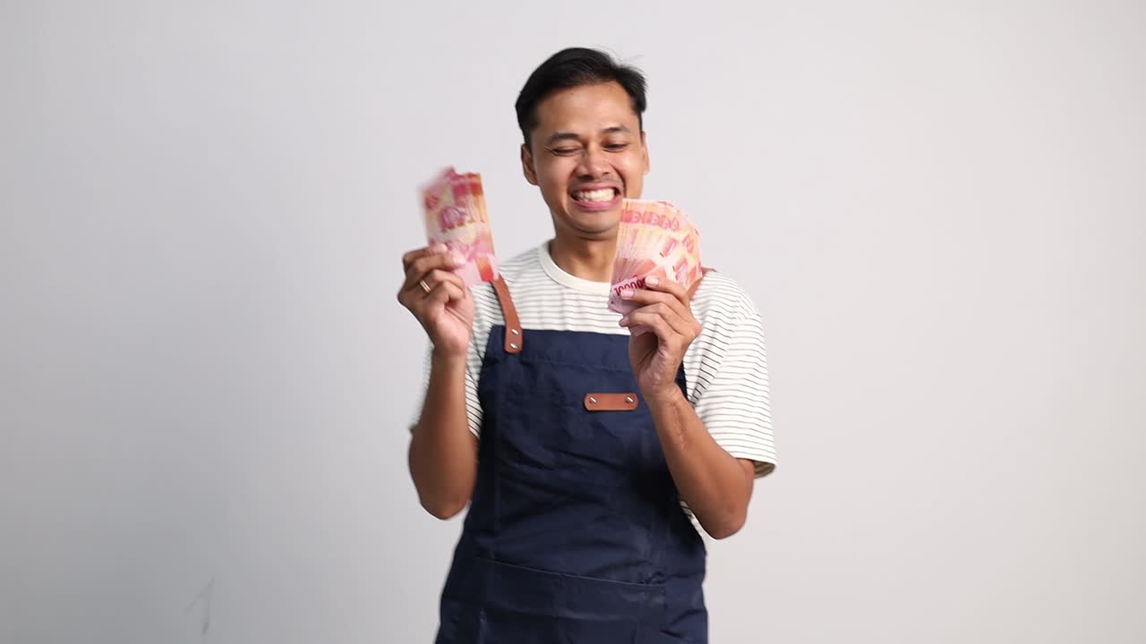 Junger asiatischer Mann mit Schürze tanzt glücklich, während er Rupiah-Banknoten hält, Gewinn oder Gehalt aus dem Geschäft