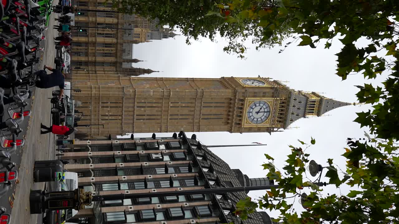 Cyclists pass London's Big Ben in slow motion with green leaves framing Elizabeth Tower. Vertical