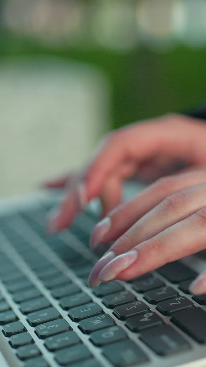 Close up of lady with polished nails typing on laptop in outdoor park setting wearing formal blazer seated on bench with blurred green trees in background