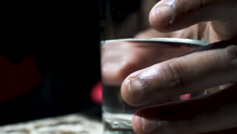 un primer plano de la mano de un hombre étnico sosteniendo un vaso lleno de un líquido claro, el hombre inquieto girando el vaso sobre la mesa con los dedos
