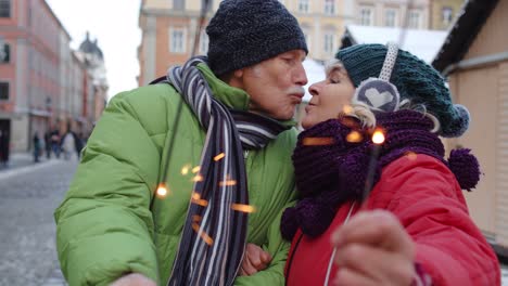 senior couple holding sparklers bengal lights enjoying christmas eve, making kiss in winter city