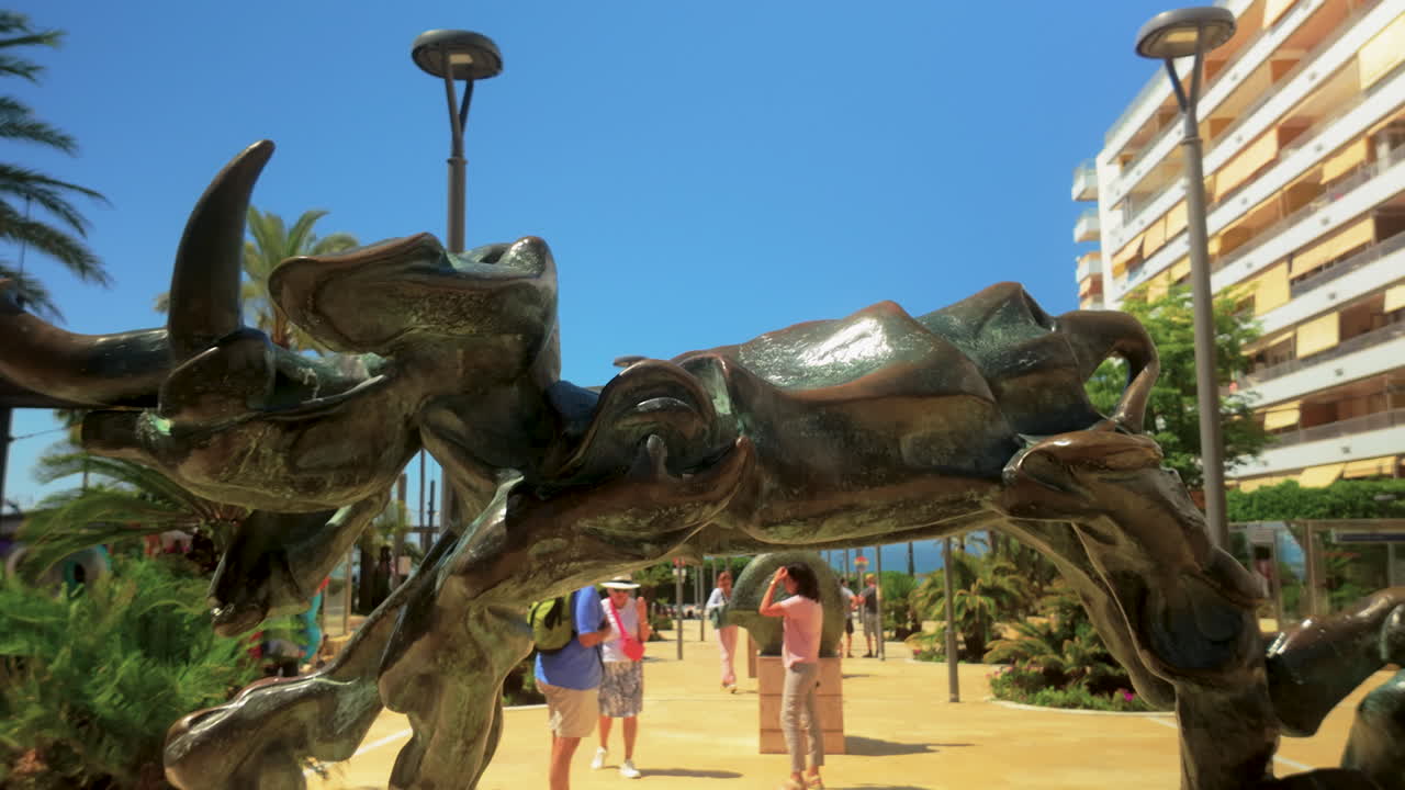 A modern bronze sculpture in a public space. The abstract form of the sculpture stands against a bright blue sky, with buildings in the background