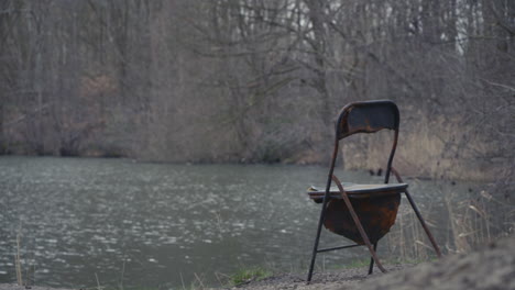 Worn-and-rusty-chair-at-a-waterfront,-background-trees-in-soft-focus