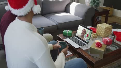 Padre-E-Hijo-Birraciales-Usando-Una-Computadora-Portátil-Para-Una-Videollamada-Navideña-Con-Un-Hombre-Sonriente-En-La-Pantalla