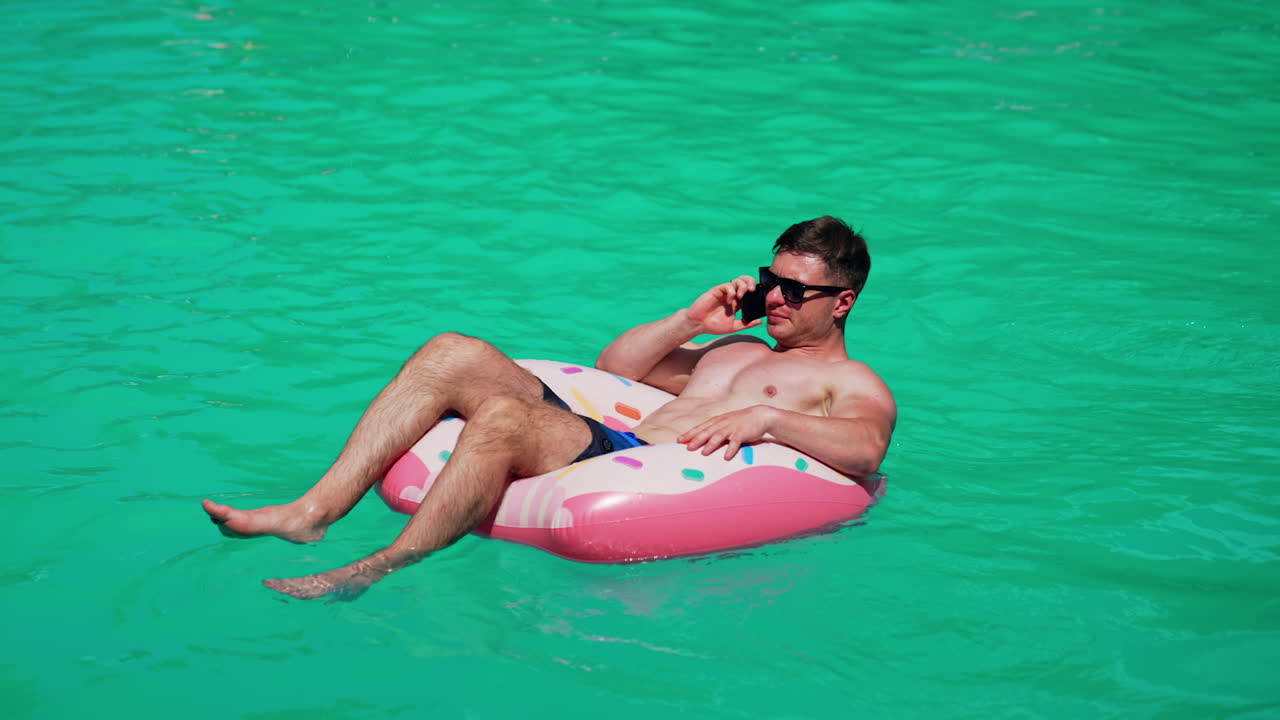 Sunbathing Caucasian man sitting in the inflatable circle talking on the phone. Male wearing sunglasses resting outdoors.