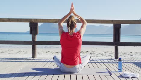 Video-of-rear-view-of-caucasian-man-with-dreadlocks-practicing-yoga-sitting-by-beach-in-sun