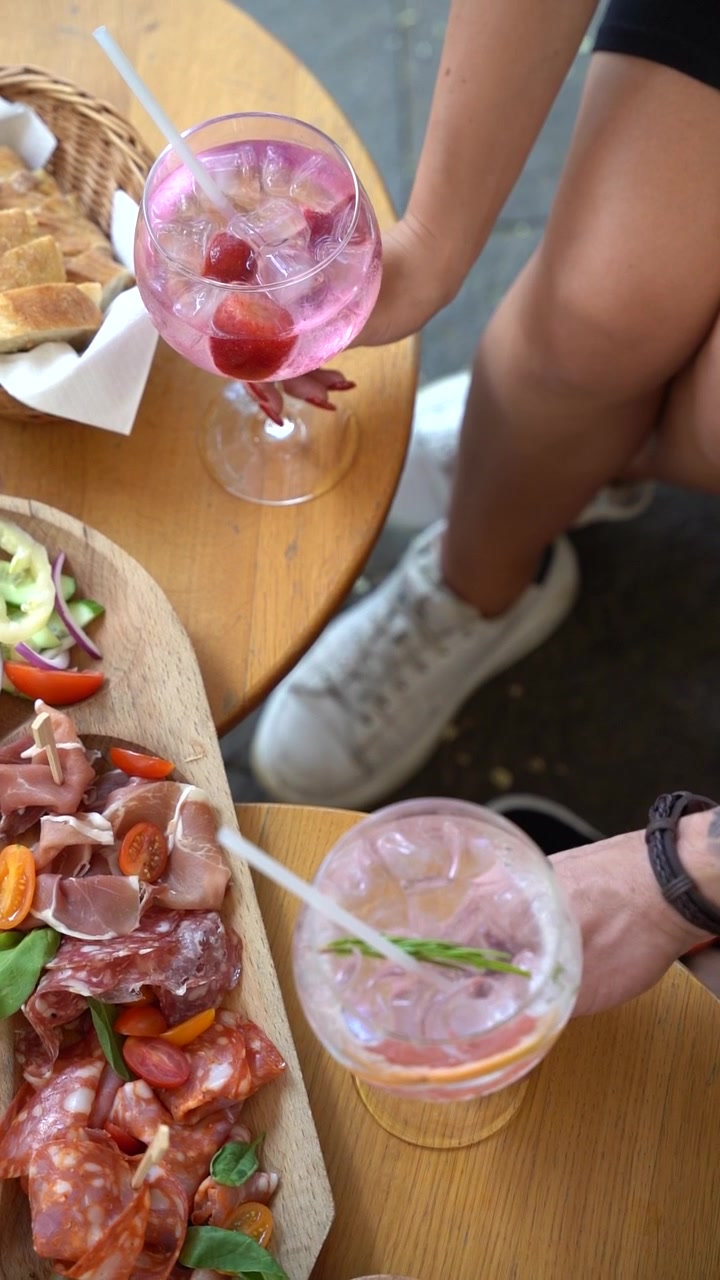 Dos manos sosteniendo un vaso de cóctel refrescante con hielo y una guarnición de romero. El fondo muestra una tabla de embutidos llena de embutidos, queso y verduras, colocada sobre una mesa de madera