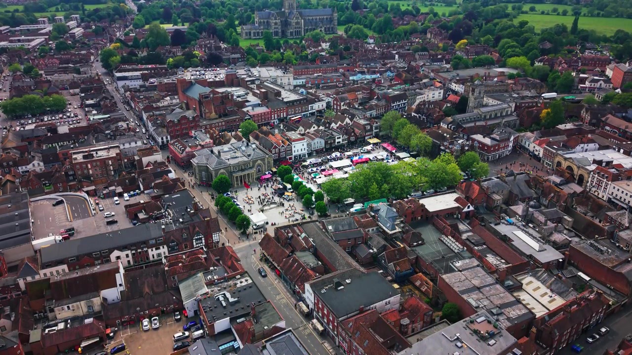 Droneopptak som ser over Salisbury sentrum med markedsboder på Market Square, folkemengder, historiske tak, veier, trær og Salisburykatedralen i det fjerne