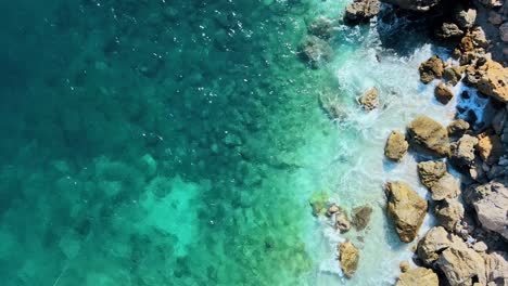 El-Agua-Clara-Del-Océano-Brilla-Mientras-Se-Estrella-En-La-Costa-Rocosa,-Fondo-Natural-Drone-De-Arriba-Hacia-Abajo