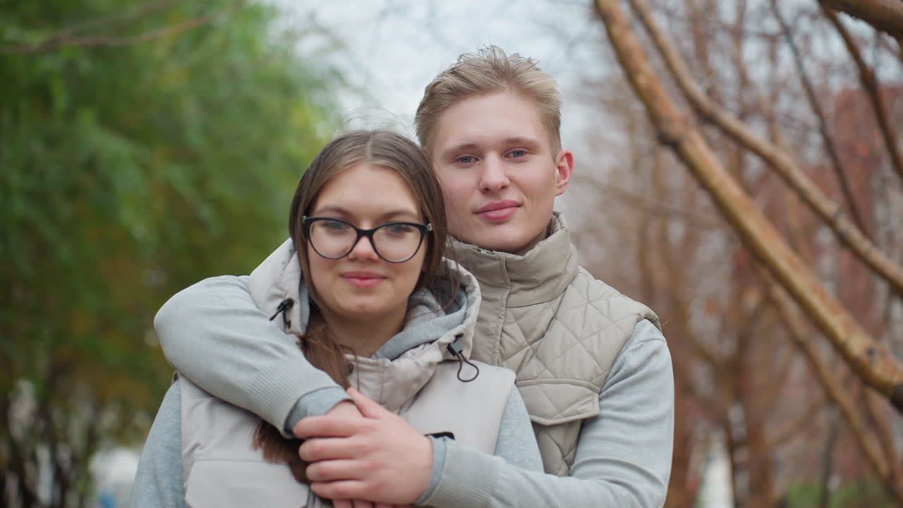 Young couple standing close together as man gently places hand across wife shoulder, both dressed in matching outfits, with tree branches swaying in wind in background