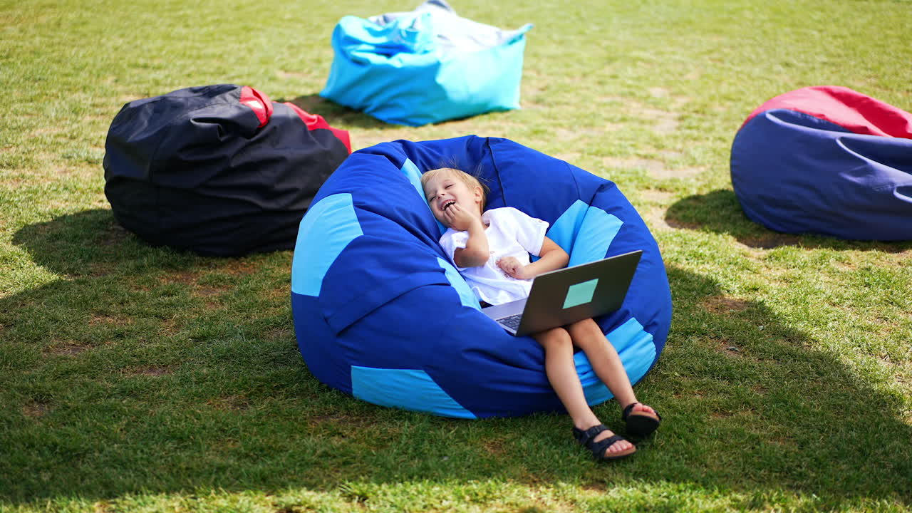 Cute boy in white shirt sits in bean bag chair with laptop on his knees. Kid watched video and laughs leaning back in chair.