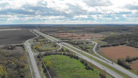 Imágenes-Aéreas-En-Las-Afueras-De-Ottawa,-Ontario,-De-Vehículos-En-Una-Carretera-Muy-Transitada-Con-Pasos-Elevados-Y-Subterráneos-Y-Líneas-Hidroeléctricas-Que-Se-Cruzan-Con-Bosques-Otoñales-Que-Rodean-Todo-Y-Campos.