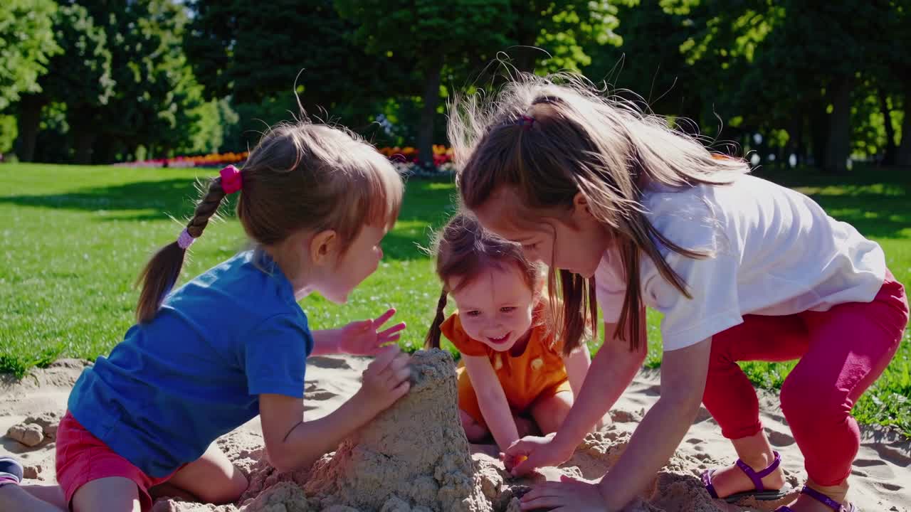 Vídeo em ângulo baixo captura três crianças construindo alegremente um castelo de areia em um parque ensolarado.